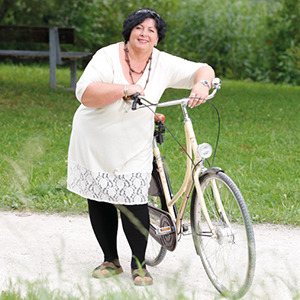 Eine mollige Frau in einem weißen Kleid und schwarzen Strümpfen lehnt an ein Fahrrad und lacht in die Kamera. Im Hintergrund ist Rasen zu sehen.