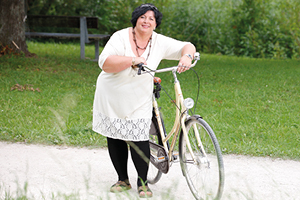 Eine mollige Frau in einem weißen Kleid und schwarzen Strümpfen lehnt an ein Fahrrad und lacht in die Kamera. Im Hintergrund ist Rasen zu sehen.
