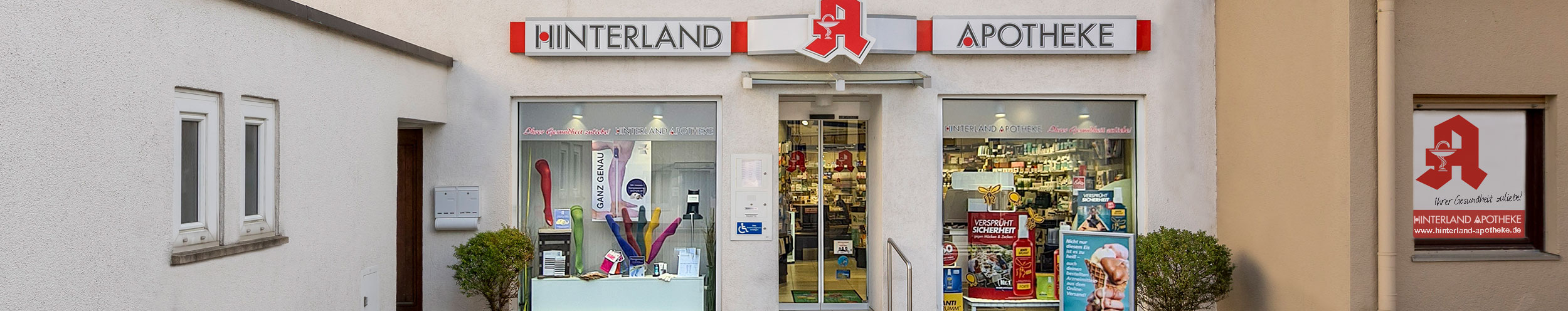 Die Außenansicht der Hinterland-Apotheke. Zu sehen ist eine Hausfront mit Schaufenstern und einem Schild mit dem Namen der Apotheke.